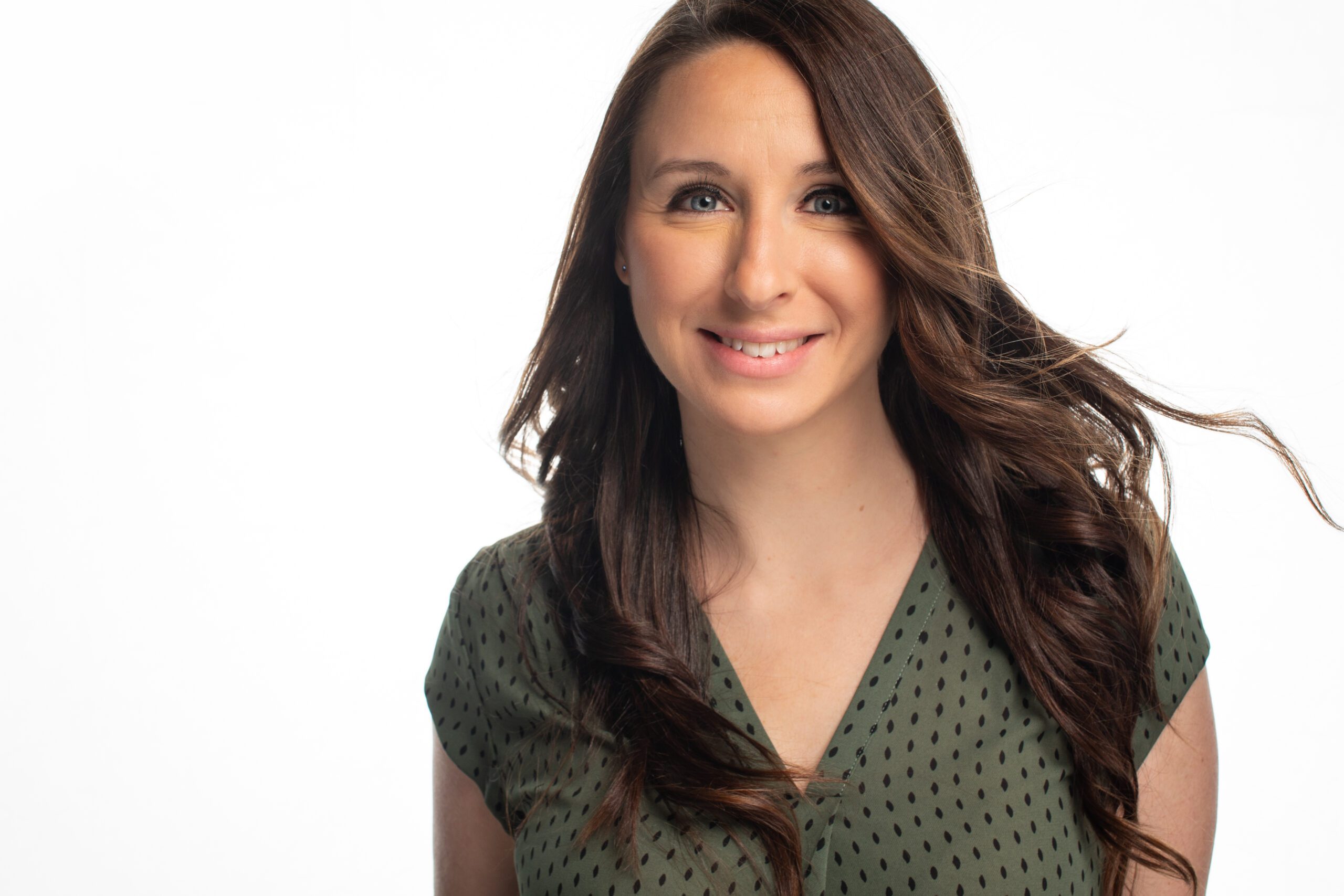 Woman with long brown hair smiling, wearing a green patterned top, against a white background.