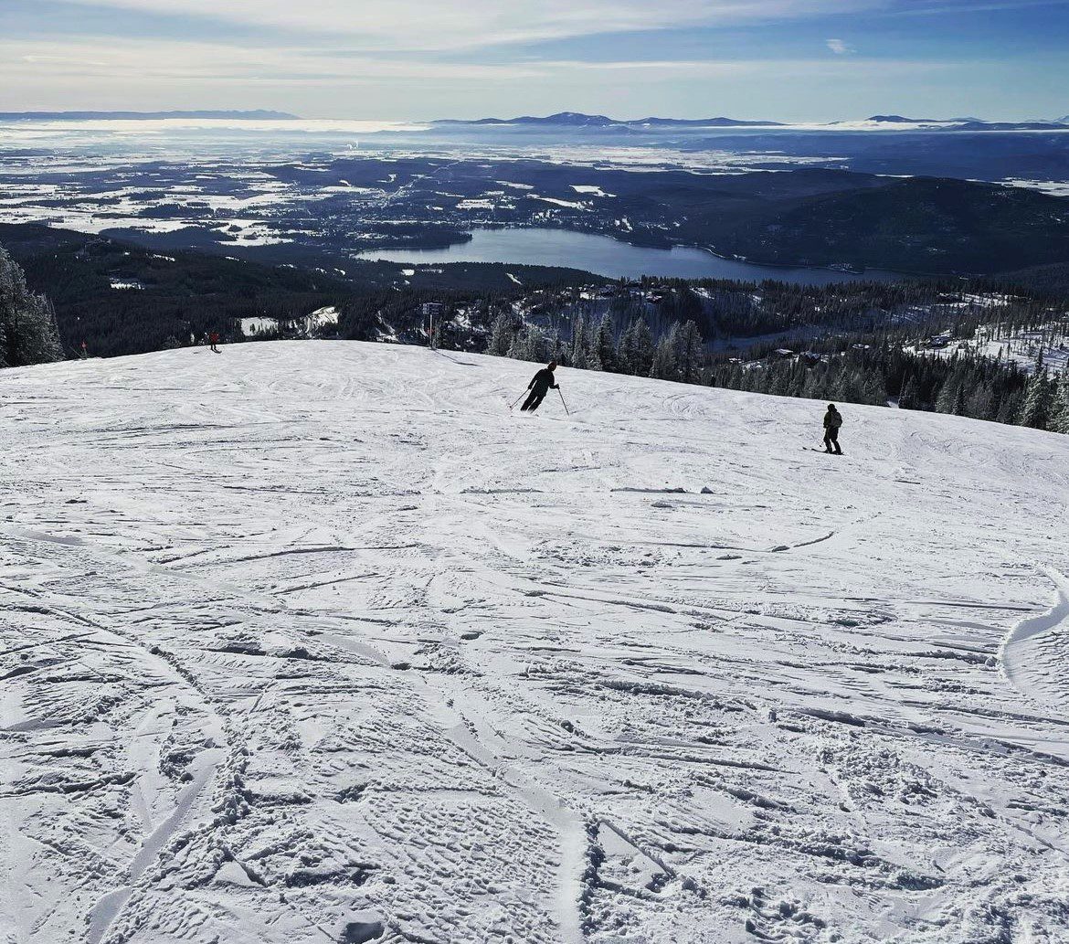 Three people skiing down a snowy slope with a panoramic view of lakes, forests, and distant mountains under a clear blue sky.